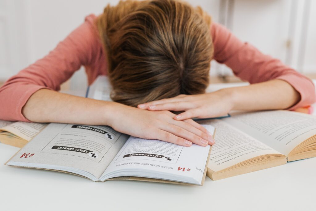 A student overwhelmed by studying falls asleep on open books at a desk, representing academic fatigue.